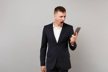 Tahriş olmuş iş adamı klasik siyah elbiseli çığlıklar, cep telefonundan arıyorum elinde, gri arka plan üzerinde yapmak video seslenmek izole. Başarı kariyer servet iş kavramı. Kopyalama alanı alay