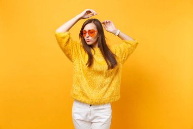 Kürk kazak, beyaz pantolon ve kalp turuncu parlak sarı arka plan üzerinde gözlük el kadar yükselen izole rahat genç kadın portresi. İnsanlar samimi duygular, yaşam kavramı. Reklam alanı