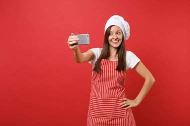 Ev hanımı kadın şef aşçı ya da baker izole kırmızı duvar arka plan üzerinde çizgili önlük, beyaz tişört, toque şefler şapkalı. Eğlence gülümseyen kadın selfie cep telefonu üzerinde atış yapıyor. Kopyalama alanı kavramı alay