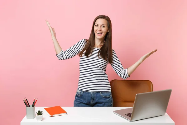 Young cheerful woman spreading hands work, standing near white desk with contemporary pc laptop isolated on pastel pink background. Achievement business career concept. Copy space for advertisement