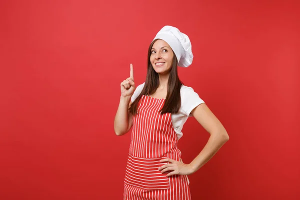 Housewife female chef cook or baker in striped apron, white t-shirt, toque chefs hat isolated on red wall background. Beautiful housekeeper woman pointing index finger up. Mock up copy space concept