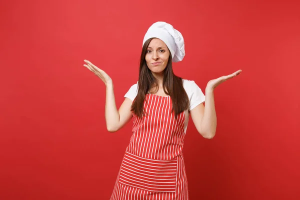 Housewife female chef cook or baker in striped apron, white t-shirt, toque chefs hat isolated on red wall background. Beautiful perplexed housekeeper woman spreading hands. Mock up copy space concept