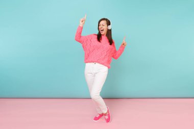 Tam uzunlukta örme gül kazak, beyaz pantolon, parlak pembe mavi pastel duvar arka planda Studio izole kulaklık mutlu genç kadın portresi. Moda yaşam tarzı kavramı. Kopyalama alanı alay