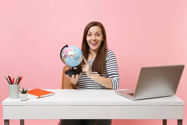 Amazed woman holding world globe and planning vacation while sit and work at white desk with contemporary pc laptop isolated on pastel pink background. Achievement business career concept. Copy space