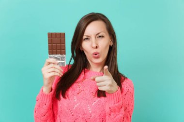 Örgü pembe kazaklı mavi duvar arka planda, stüdyo portre izole kamera tutun el çikolata bar işaret parmağı işaret çarpıcı genç kadın. İnsanlar yaşam tarzı kavramı. Kopyalama alanı alay
