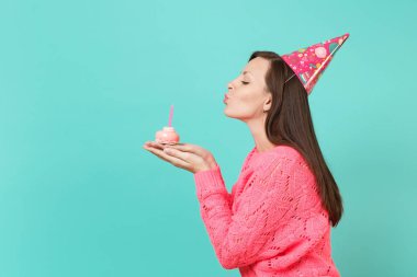 Örgü pembe kazaklı, pasta elinde mum dışarı üfleme Doğum günü şapkası genç kadın yan görünüm mavi turkuaz renkli stüdyo portre üzerinde izole. İnsanlar yaşam tarzı kavramı. Kopyalama alanı alay