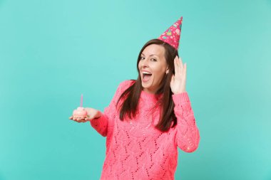 Örgü pembe kazaklı neşeli genç kadın, Doğum günü şapkası kulak misafiri, elinde mavi duvar arka plan üzerinde izole mum ile pasta tutan işitme jest. İnsanlar yaşam tarzı kavramı. Kopyalama alanı alay