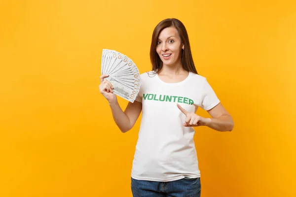 Woman in white t-shirt with written inscription green title volunteer hold lots dollars banknotes, cash money isolated on yellow background. Voluntary free assistance help, charity grace work concept