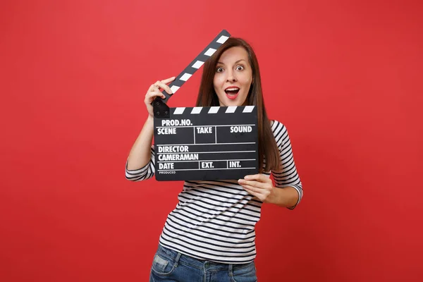 Excited young woman keeping mouth wide open looking surprised holding classic black film making clapperboard isolated on red background. People sincere emotions, lifestyle concept. Mock up copy space