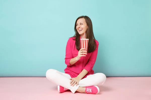 Full length portrait woman in rose shirt blouse, white pants sitting on floor hold cup of soda isolated on pink blue pastel wall background studio. Fashion lifestyle concept. Mock up copy space