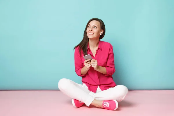 Full length portrait woman in rose shirt blouse, white pants sitting on floor with earphones isolated on bright pink blue pastel wall background studio. Fashion lifestyle concept. Mock up copy space