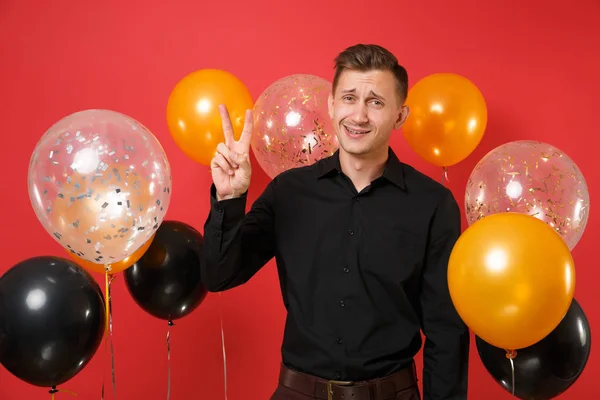 Smirked young man in black classic shirt celebrating, showing victory sign on red background air balloons. Valentine\'s, International Women\'s Day Happy New Year, birthday mockup holiday party concept