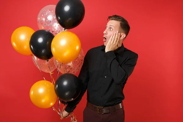 Excited young man in black classic shirt holding air balloons, looking aside, putting hand on face isolated on red background. St. Valentine\'s Day Happy New Year birthday mockup holiday party concept