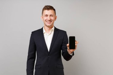 Klasik siyah takım elbise ve gömlek gri duvar arka plan üzerinde izole boş boş ekran ile cep telefonu holding neşeli genç iş adamı. Başarı kariyer servet iş kavramı. Kopyalama alanı alay