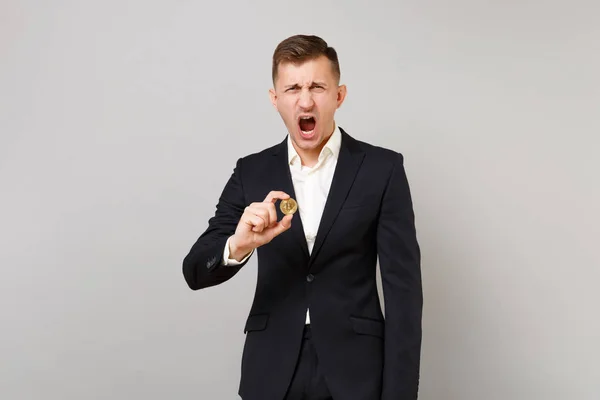 Irritated young business man in classic black suit, shirt holding bitcoin, future currency, screaming isolated on grey wall background. Achievement career wealth business concept. Mock up copy space