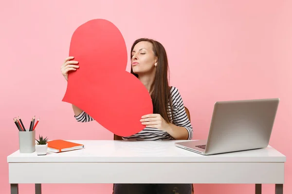 Tender woman blowing lips sending air kiss holding red empty blank heart sit and work at white desk with pc laptop isolated on pastel pink background. Achievement business career concept. Copy space