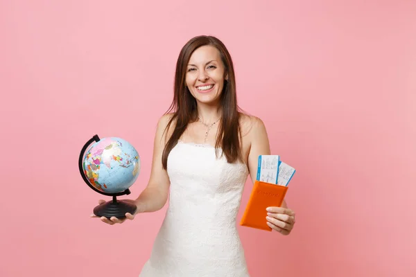 Happy bride woman in white wedding dress holding world globe, passport boarding pass ticket, going abroad for honeymoon, vacation isolated on pastel pink background. Wedding celebration. Copy space