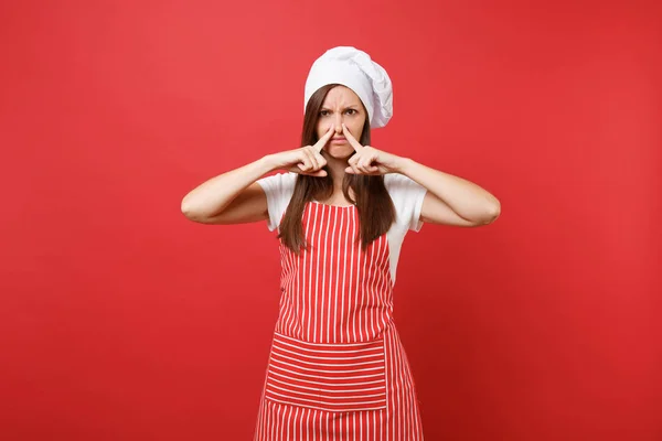 Housewife female chef cook or baker in striped apron, white t-shirt, toque chefs hat isolated on red wall background. Housekeeper woman close nose with hand because smell. Mock up copy space concept