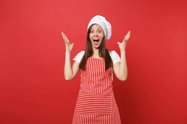 Housewife female chef cook baker in striped apron, white t-shirt, toque chefs hat isolated on red wall background. Shocked mad crazy fun housekeeper woman spreading hands. Mock up copy space concept