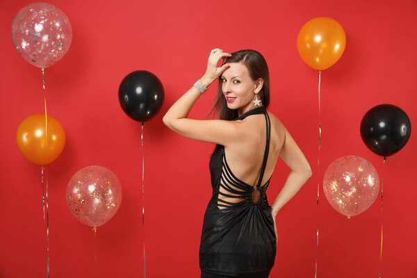 Back rear view of beautiful girl in little black dress looking back celebrating on bright red background air balloons. International Women's Day, Happy New Year, birthday mockup holiday party concept