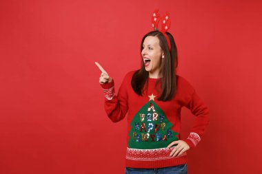 Dekoratif geyik boynuzları işaret parmağı bir yana, ağız açık tutarak işaret eden kırmızı zemin üzerine izole eğlenceli neşeli Noel Baba kız. Mutlu yeni yıl 2019 kutlama tatil parti kavramı. Kopyalama alanı alay