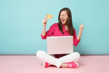 Tam uzunlukta gül gömlekli genç kadın portresi, beyaz pantolon katta oturan pembe mavi pastel duvar arka plan studio izole kredi kartı pc tutun. Moda yaşam tarzı kavramı. Kopyalama alanı alay