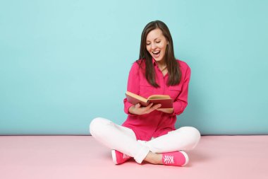Tam uzunlukta gül gömlek bluz gülümseyen kadın portresi, beyaz pantolon pembe mavi pastel duvar arka plan studio izole kitap okumak katta oturmak. Moda yaşam tarzı kavramı. Kopyalama alanı alay
