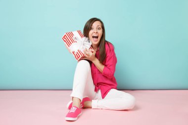 Tam uzunlukta gül gömlek bluz, kadın portre, beyaz pantolon katta oturan mevcut kutusu pembe mavi pastel duvar arka plan studio izole tutun. Moda yaşam tarzı kavramı. Kopyalama alanı alay.