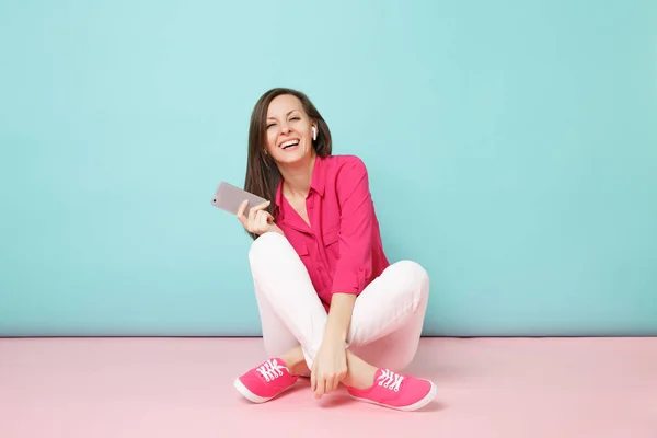 Full length portrait woman in rose shirt blouse, white pants sitting on floor with earphones isolated on bright pink blue pastel wall background studio. Fashion lifestyle concept. Mock up copy space