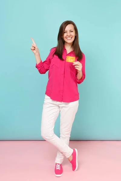 穿着玫瑰衬衫衬衫的年轻女子的全长肖像 白色裤子持有信用卡隔离在明亮的粉红色蓝色柔和墙背景工作室 时尚生活方式的概念 模拟复制空间 — 图库照片