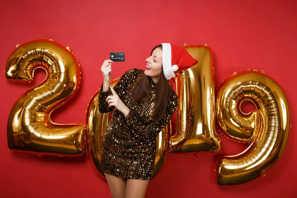 Merry Santa girl in shiny glitter dress, Christmas hat hold credit card isolated on bright red wall background, golden numbers air balloons studio portrait. Happy New Year 2019 holiday party concept