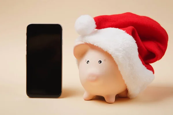 Piggy money bank with Christmas hat, mobile phone with blank empty screen isolated on beige background. Money accumulation investment, banking services, wealth concept. Copy space advertising mock up