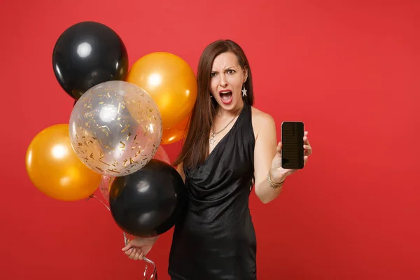Angry young woman in black dress screaming celebrating holding mobile phone with blank black empty screen air balloons isolated on red background. Happy New Year birthday mockup holiday party concept