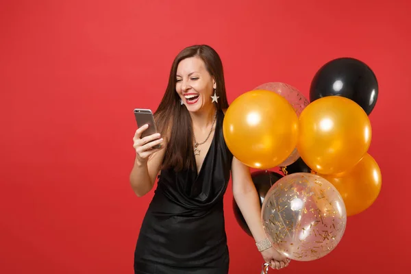 Laughing young woman in little black dress holding air balloons and using mobile phone while celebrating isolated on red background. Women\'s Day, Happy New Year, birthday mockup holiday party concept