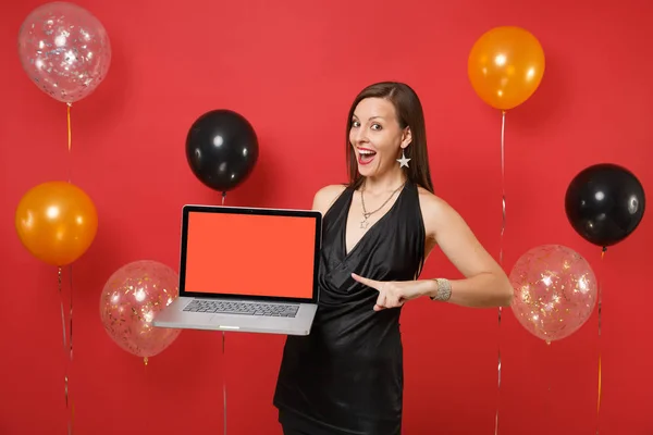 Excited woman in black dress pointing index finger on laptop pc computer with blank black empty screen on bright red background air balloons. Happy New Year, birthday mockup holiday party concept