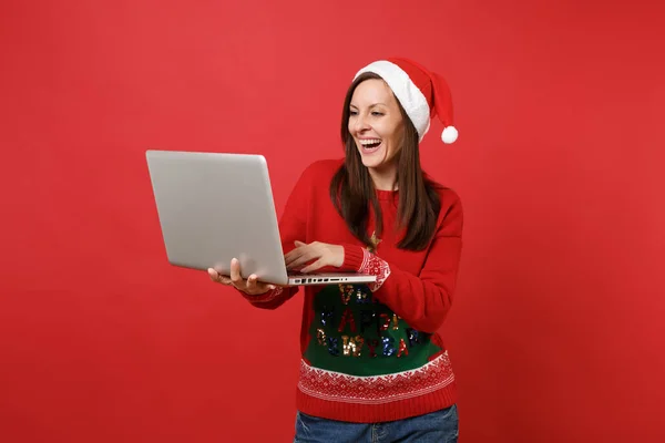 Laughing young Santa girl in knitted sweater, Christmas hat working, typing on laptop pc computer isolated on red background. Happy New Year 2019 celebration holiday party concept. Mock up copy space