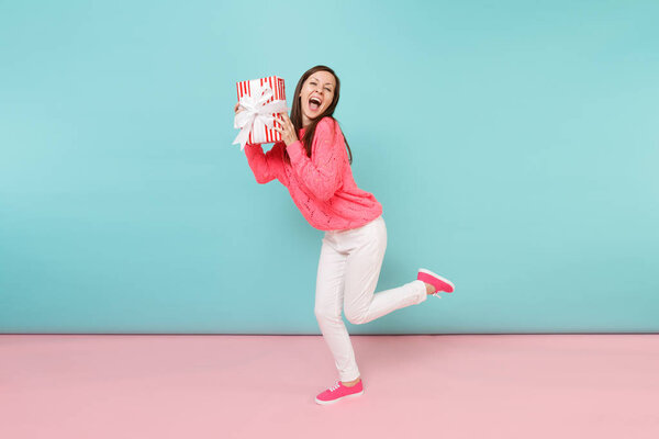 Full length portrait of woman in knitted rose sweater, white pants posing holding gift box isolated on bright pink blue pastel wall background in studio. Fashion lifestyle concept. Mock up copy space