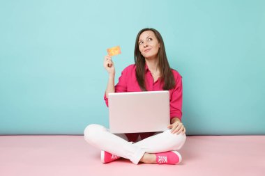 Tam uzunlukta gül gömlekli genç kadın portresi, beyaz pantolon katta oturan pembe mavi pastel duvar arka plan studio izole kredi kartı pc tutun. Moda yaşam tarzı kavramı. Kopyalama alanı alay