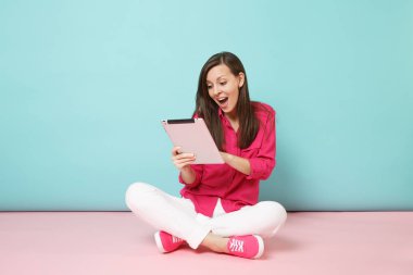 Tam uzunlukta gül gömlek bluz, kadın portre, beyaz pantolon katta oturan pembe mavi pastel duvar arka plan studio izole tablet pc tutun. Moda yaşam tarzı kavramı. Kopyalama alanı alay.