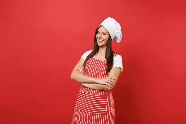 Housewife female chef cook or baker in striped apron, white t-shirt, toque chefs hat isolated on red wall background. Pretty young housekeeper woman holding hands crossed. Mock up copy space concept