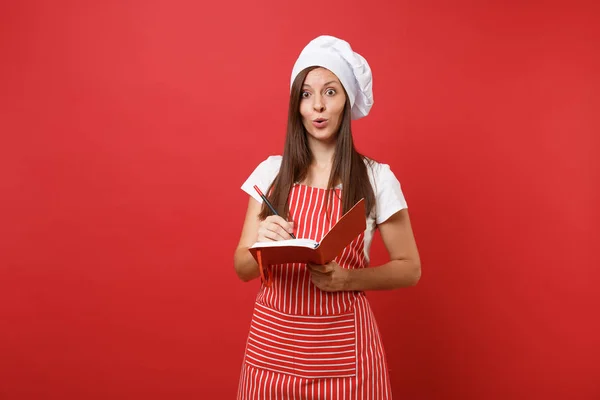 Housewife female chef cook or baker in striped apron white t-shirt toque chefs hat isolated on red wall background. Smiling woman hold notepad recipe cooking book and pen. Mock up copy space concept