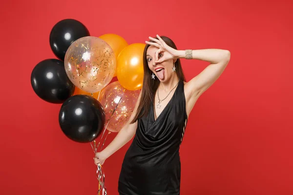 Funny crazy woman in black dress showing tongue OK sign near eyes holding air balloons celebrating isolated on red background. St. Valentine\'s Day Happy New Year birthday mockup holiday party concept