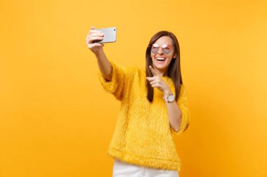 Gülen genç kadın kazak kalp bardaklarda alarak selfie cep telefonu işaret parmağı parlak sarı arka plan üzerinde izole atış yapıyor. İnsanlar samimi duygular, yaşam tarzı. Reklam alanı