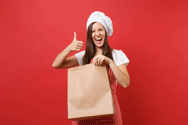 Housewife female chef cook baker in striped apron t-shirt toque chefs hat isolated on red wall background.Woman hold in hand brown clear blank craft paper bag takeaway Mock up copy space concept