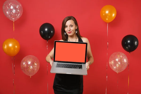 Beautiful young woman in black dress celebrating holding laptop pc computer with blank black empty screen on bright red background air balloons. Happy New Year, birthday mockup holiday party concept