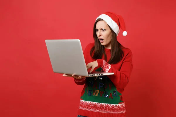 Concerned shocked young Santa girl in Christmas hat working, typing on laptop pc computer isolated on bright red background. Happy New Year 2019 celebration holiday party concept. Mock up copy space