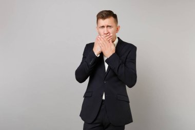 Portresi üzgün genç iş adamı klasik siyah takım elbise, gömlek gri duvar arka planda Studio izole ellerle ağız kapsayan. Başarı kariyer servet iş kavramı. Kopyalama alanı alay