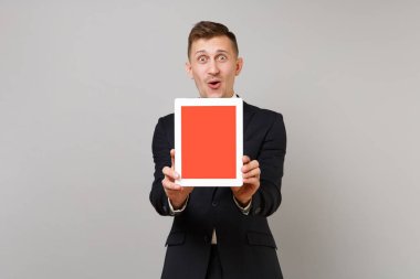 Sürpriz genç iş adamı klasik siyah takım elbise gömlek gri arka plan üzerinde izole boş boş ekran ile tablet pc bilgisayar tutarak. Başarı kariyer servet iş kavramı. Kopyalama alanı alay