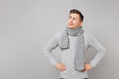 Şaşkın genç adam gri süveterli eşarp silah bir kenara gri arka plan üzerinde izole bel arayan akimbo ile. Sağlıklı moda yaşam tarzı insanların samimi duygular soğuk sezon kavramı. Kopyalama alanı alay