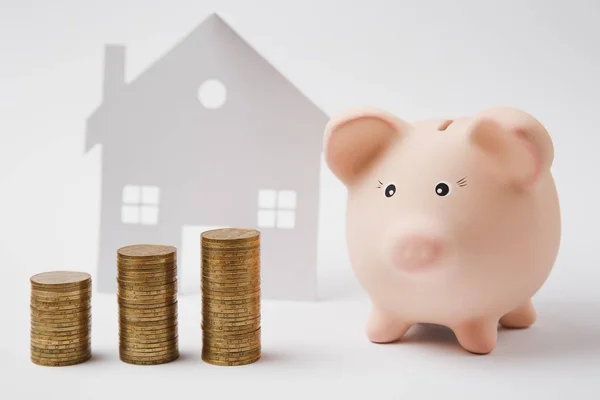 Close up of pink piggy money bank stacks of golden coins on white wall background with house. Money accumulation investment banking or business services wealth concept. Copy space advertising mock up